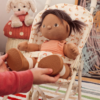 Doll with dark hair and brown shoes sitting in a baby bouncer, held by a hand.