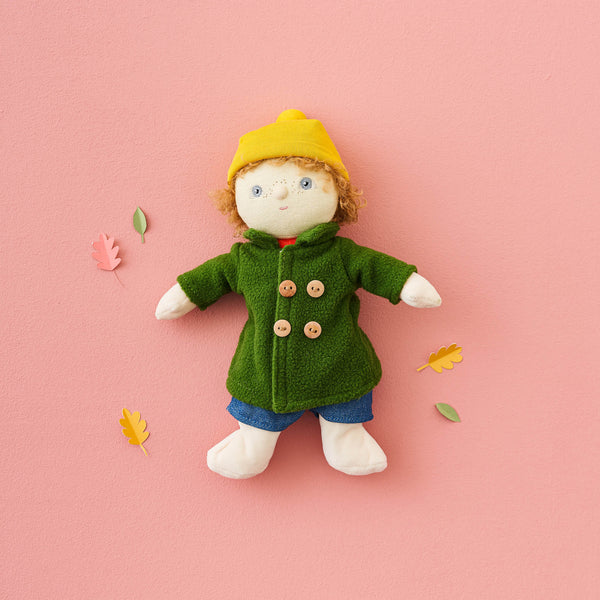 Toy figure wearing a green coat and yellow hat on a pink background with autumn leaves.