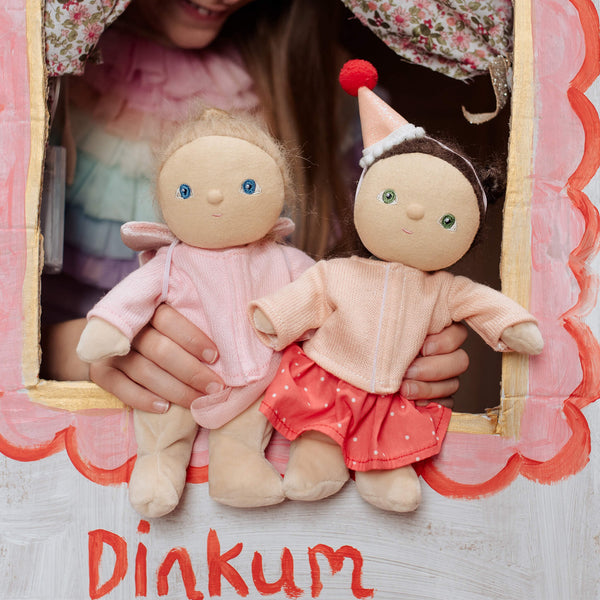 Two dolls held by a person with a decorative frame, featuring the brand 'Dinkum'.
