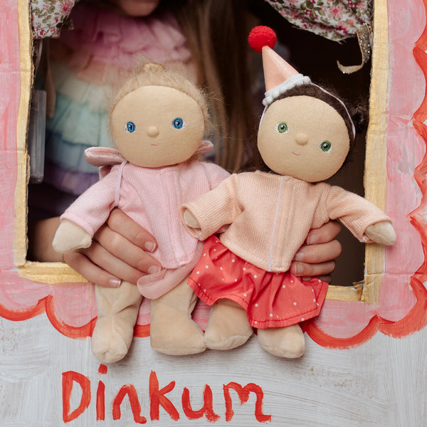 Two dolls held by a person with 'Dinkum' branding on a wooden surface.